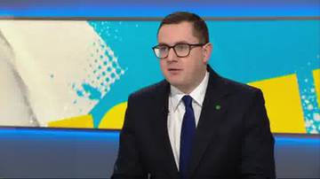 Mężczyzna w okularach i ciemnym garniturze siedzi na tle niebiesko-żółtego ekranu z abstrakcyjnymi wzorami.