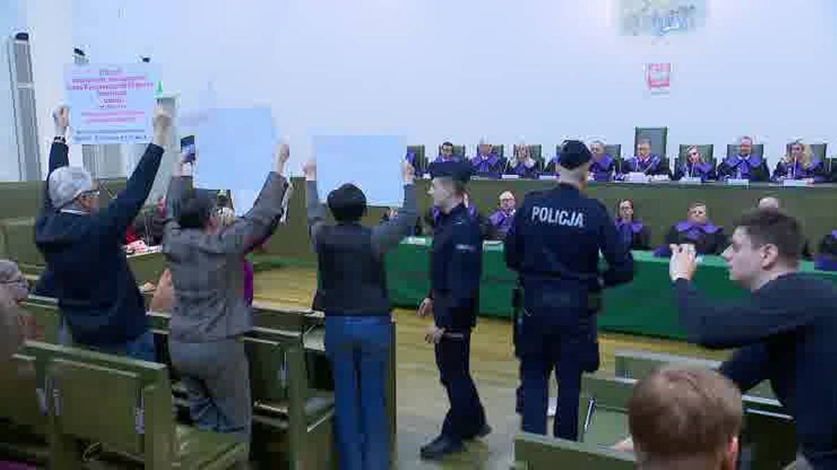 Grupa osób protestujących trzymających transparenty w sali sądowej, w której interweniuje policja.