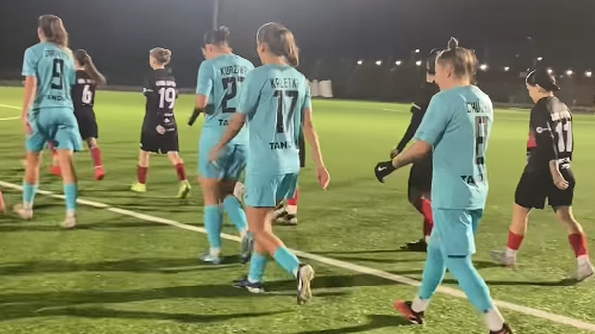 Grupa piłkarek w jasnoniebieskich strojach sportowych schodzi z zielonego boiska piłkarskiego. Widoczne są numery na koszulkach, m.in. 9, 27, 17. W tle znajdują się inne zawodniczki w czarnych strojach.