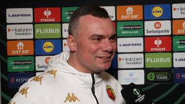 Adrian Siemieniec, trener, uśmiechnięty, w bluzie z kapturem, na tle paneli sponsorów.