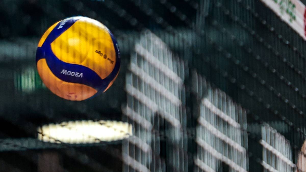 Siatkarz w czarnej koszulce z ż&oacute;łtymi elementami odbija piłkę nad siatką, w tle widzowie na trybunach.