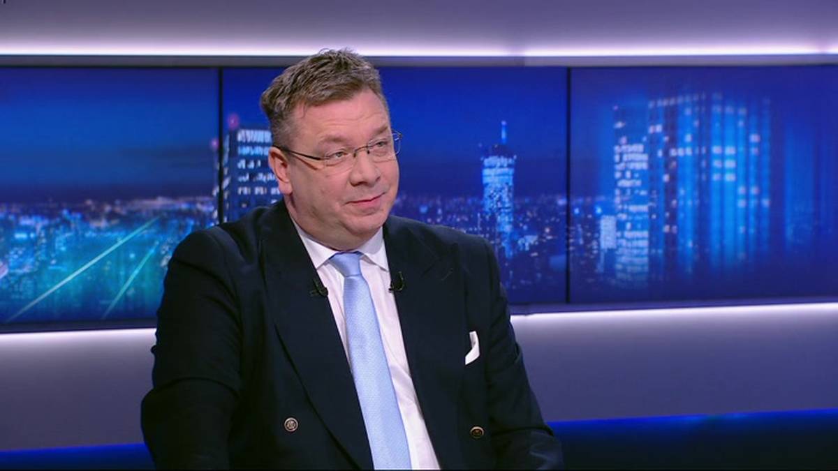 Mężczyzna w średnim wieku, ubrany w ciemny garnitur i jasny krawat, siedzi przed ekranami wyświetlającymi nocną panoramę miasta.