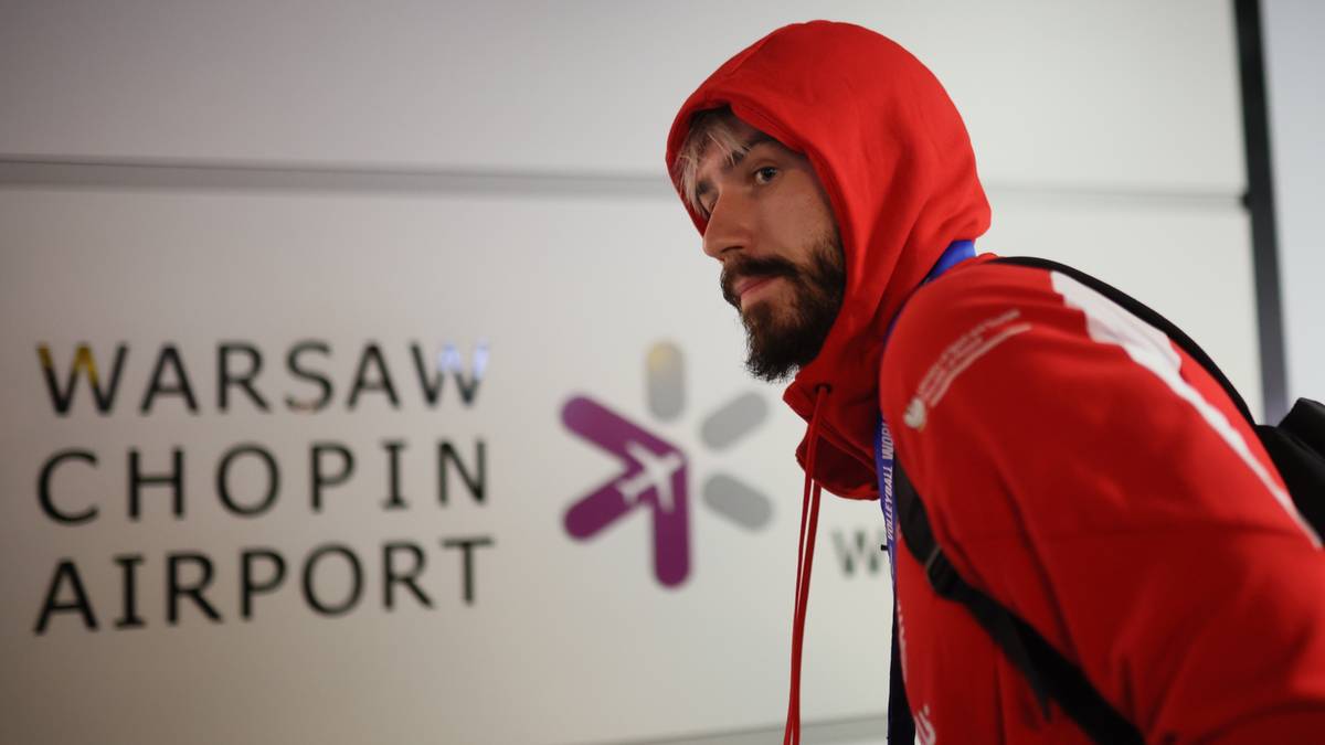 Mężczyzna w czerwonej bluzie z kapturem stoi przed napisem "WARSAW CHOPIN AIRPORT" i logo lotniska.