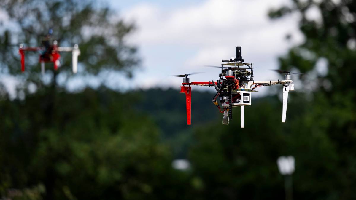 Dwa drony unoszą się w powietrzu w trakcie wyścigu. Jeden dron, znajdujący się na pierwszym planie, jest bardziej szczegółowy, z czerwonymi ramionami i białymi elementami konstrukcyjnymi. W tle widać drugi dron, częściowo zasłonięty przez drzewa. Niebo jest błękitne z białymi chmurami.