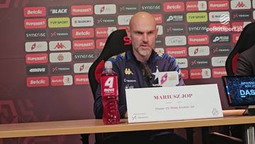 Mariusz Jop, trener Wisły Kraków, siedzi przy stole z napisem "MARIUSZ JOP" i butelką napoju.