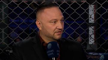 Portret Tomasza Babilońskiego, mężczyzny z kr&oacute;tkimi włosami i zarostem, w czarnej marynarce i koszuli, trzymającego mikrofon, z siatką klatki ringowej w tle.