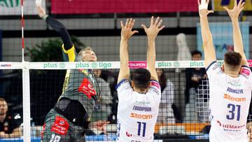 Siatkarz w ż&oacute;łto-czarnej koszulce wykonuje atak nad siatką, blokowany przez dw&oacute;ch zawodnik&oacute;w w białych koszulkach.