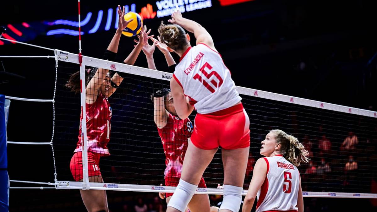 Siatkarka w białej koszulce z numerem 15 i czerwonymi spodenkami wykonuje atak nad siatką, odbijając piłkę.