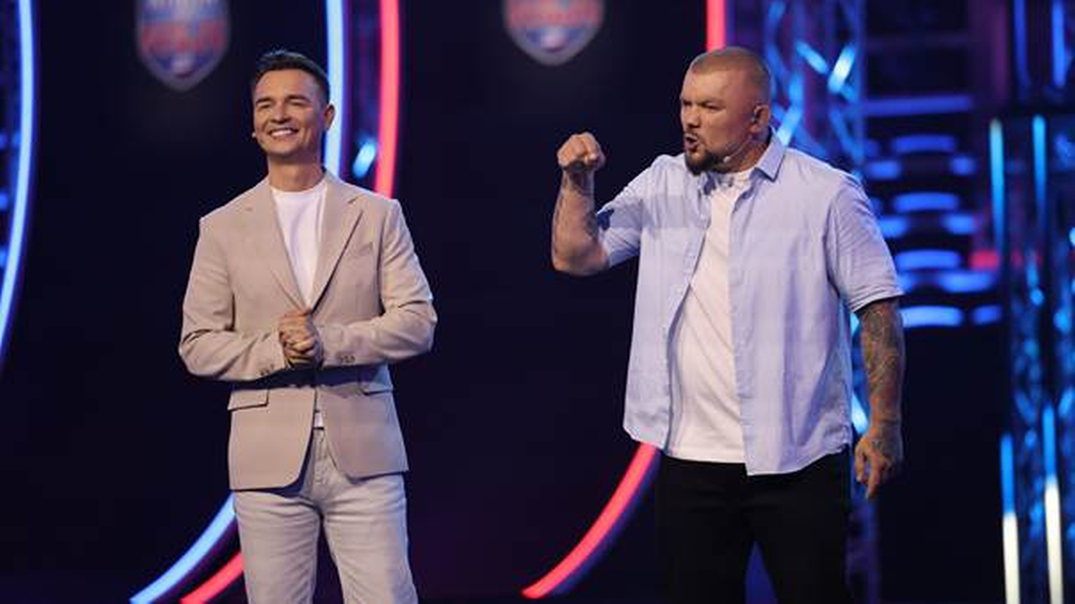 Dwóch mężczyzn stoi na scenie oświetlonej niebieskim i czerwonym światłem, z elementami przypominającymi tarcze na tle.