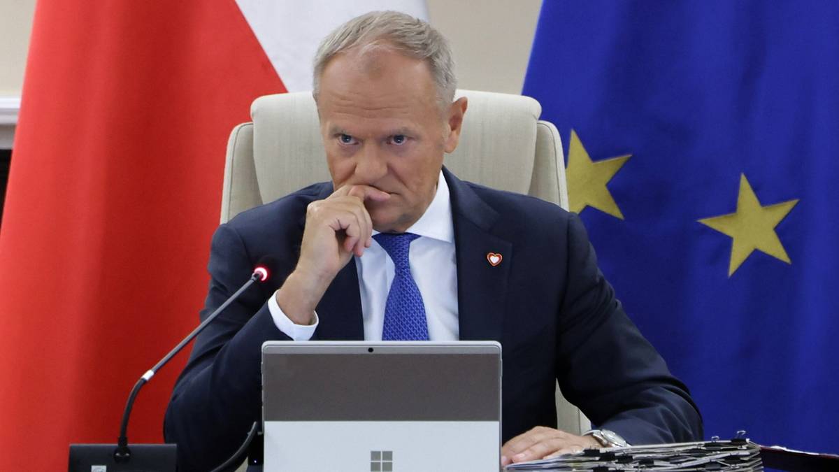 Donald Tusk siedzi przy stole, ubrany w ciemny garnitur i niebieski krawat. Jest pochylony do przodu, opierając palce prawej dłoni o brodę, z zamyśloną miną. Przed nim leży tablet. W tle widoczne są flagi Polski i Unii Europejskiej.