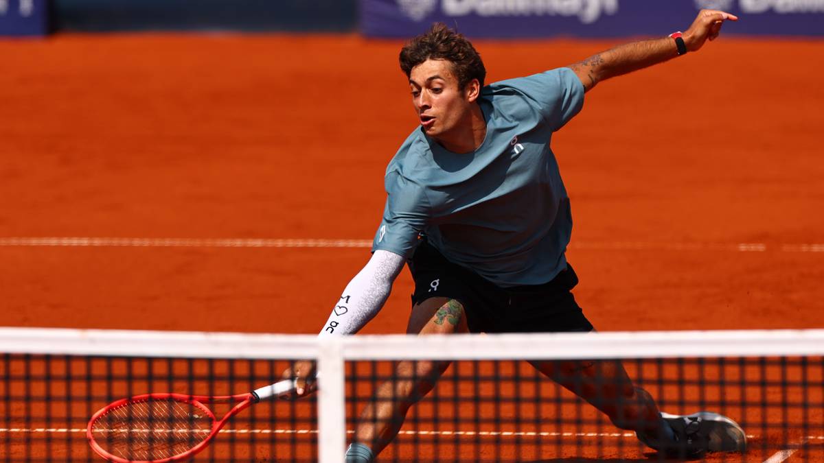 Tenisista w akcji na korcie ziemnym, odbija piłkę rakietą nad siatką.