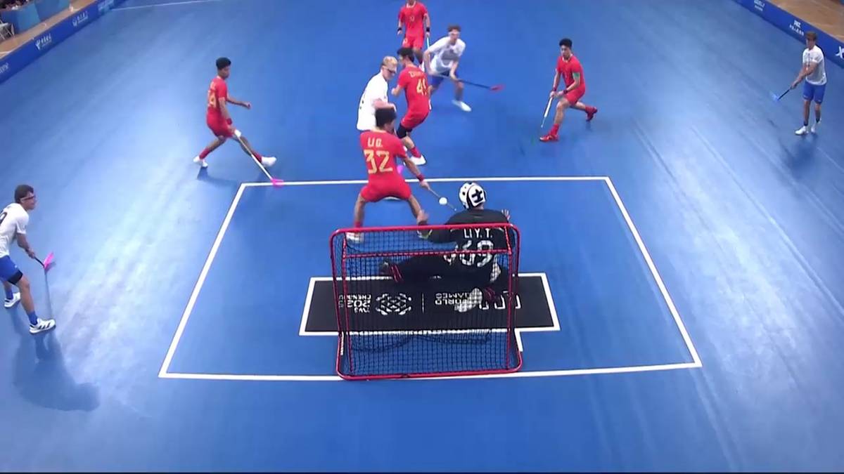 Gracze drużyny w czerwonych strojach rywalizują z drużyną w białych strojach w meczu unihokeja. Jeden z zawodników w czerwonym stroju z numerem 32 na plecach próbuje oddać strzał, podczas gdy inny zawodnik w białym stroju z numerem 32 próbuje go zablokować. Bramkarz w czarnym stroju znajduje się w bramce. Dwóch innych zawodników w czerwonych strojach biegnie w kierunku bramki. Akcja rozgrywa się na niebieskiej hali sportowej z białymi liniami wyznaczającymi boisko.