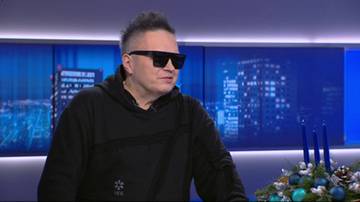 Muniek Staszczyk w czarnej bluzie z kapturem i okularach przeciwsłonecznych, siedzi przed niebieskim ekranem z widokiem na nocne miasto. Po prawej stronie kadru widoczny jest świąteczny stroik z bombkami i świecami.