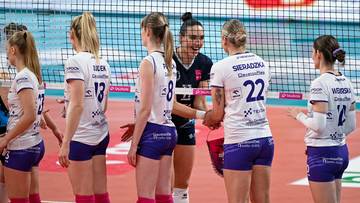 Grupa siatkarek w białych koszulkach i niebieskich spodenkach stoi na boisku siatkarskim, jedna z nich uśmiecha się do pozostałych.