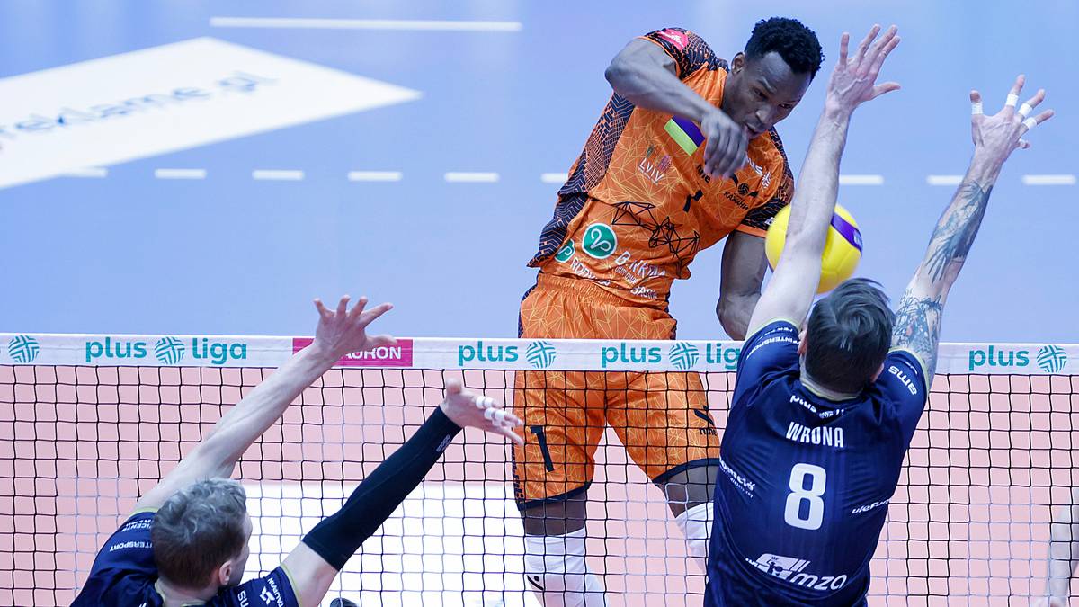 Gracz siatkówki w pomarańczowym stroju skacze do ataku, blokowany przez dwóch graczy w niebieskich strojach.