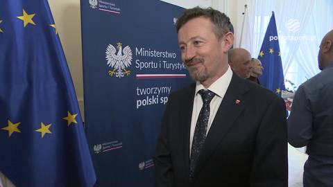 Adam Małysz, ubrany w garnitur, uśmiecha się do obiektywu. W tle znajduje się niebieska tablica z logo Ministerstwa Sportu i Turystyki oraz flagi Polski i Unii Europejskiej.