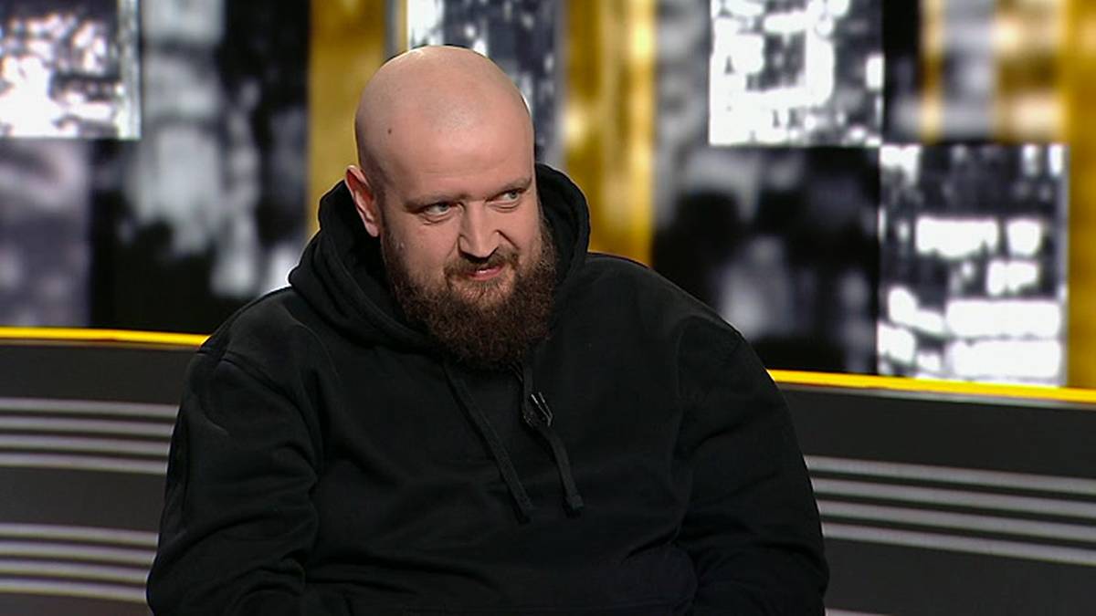 Mężczyzna z łysiną i brodą w czarnej bluzie z kapturem uśmiecha się krzywo.