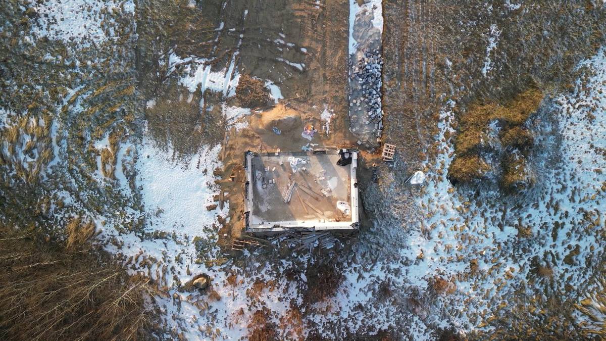 Plac budowy domu z widocznymi fundamentami i częściowo postawioną ścianą, przykrytymi plandeką, zimą.