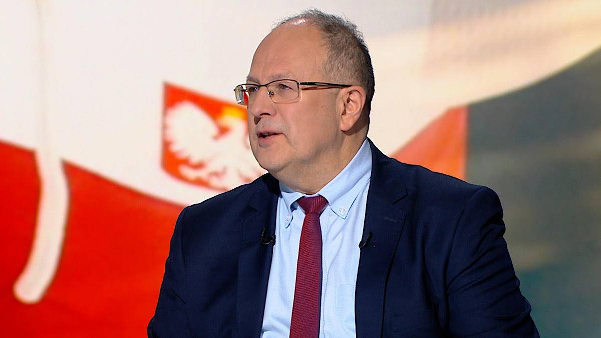 Mężczyzna w okularach i granatowym garniturze, w koszuli i krawacie, siedzi przed biało-czerwoną flagą.