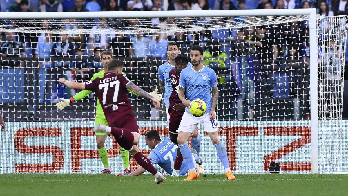 Piłkarz w bordowym stroju z numerem 77 na plecach kopie piłkę w kierunku bramki, obok niego w niebieskim stroju leży inny zawodnik, w tle bramkarz w zielonym stroju i widzowie.