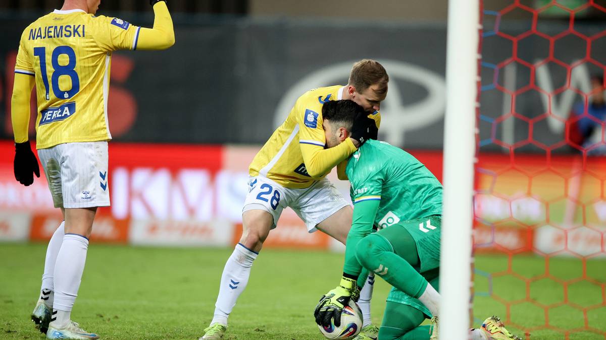 Piłkarze walczą o piłkę w polu karnym, bramkarz w zielonym stroju klęczy na murawie.