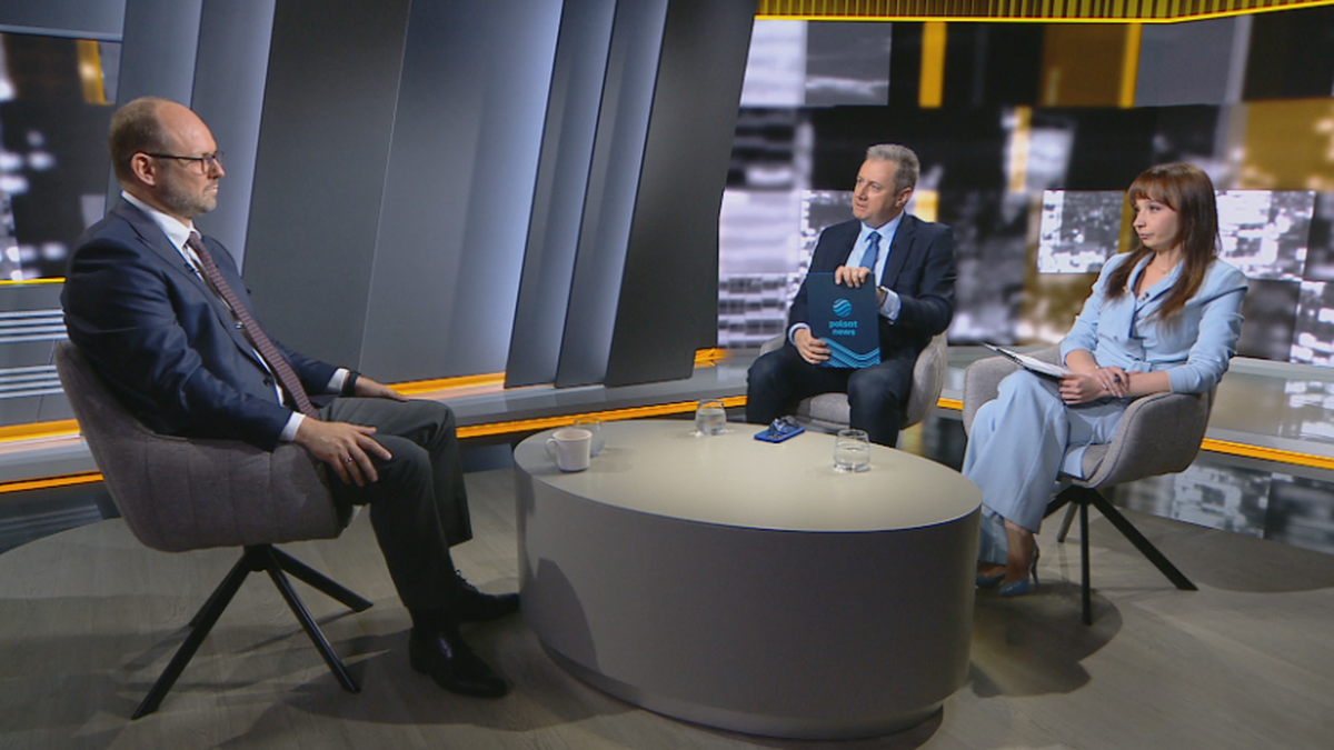 Marcin Przydacz, ubrany w garnitur i krawat, siedzi na krześle po lewej stronie. Na środku stołu znajduje się biały kubek. Po prawej stronie siedzą dwie osoby: mężczyzna w garniturze trzyma teczkę, a kobieta w jasnym kostiumie notuje coś na kartce. W tle znajduje się nowoczesne studio z geometrycznymi elementami i podświetleniem.