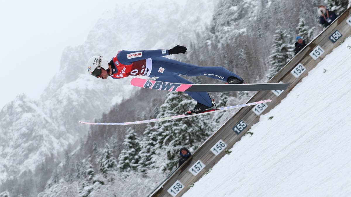 Skoczek narciarski w locie na skoczni mamuciej w zimowych warunkach, otoczony śniegiem i zaśnieżonymi g&oacute;rami.