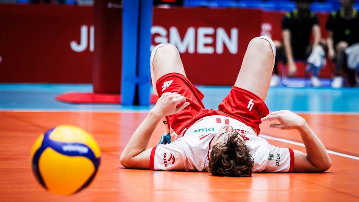 Siatkarz leżący na boisku z nogami uniesionymi do góry, w koszulce z biało-czerwonym godłem Polski.