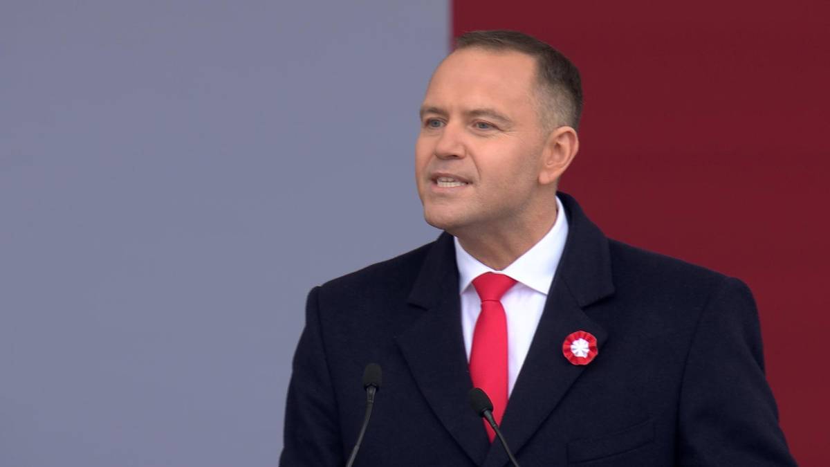 Karol Nawrocki przemawia do mikrofonu w czarnym garniturze i czerwonym krawacie.