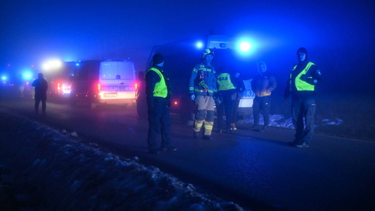 Strażacy w kamizelkach odblaskowych i pojazdy ratunkowe z włączonymi niebieskimi światłami na tle nocnego krajobrazu.