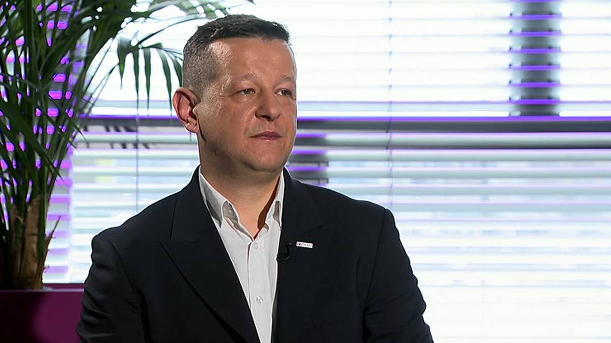 Portret mężczyzny w garniturze, siedzącego na tle żaluzji z fioletowymi pasami, obok rośliny doniczkowej.