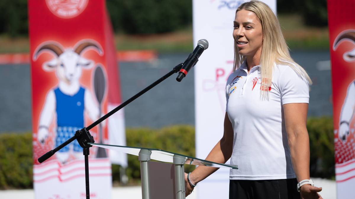 Kobieta z blond włosami przemawia do mikrofonu stojąc przy m&oacute;wnicy. Za nią widoczne są czerwone banery z wizerunkami k&oacute;z.