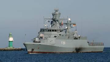 Szara korweta wojenna z numerem burtowym F 261 na wodzie obok falochronu z latarnią morską.