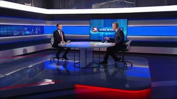 Dw&oacute;ch mężczyzn w eleganckich garniturach siedzi przy okrągłym stole w nowoczesnym studiu telewizyjnym, przed ekranem z logo programu "Gość Wydarzeń".