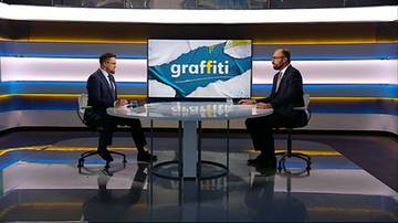Dw&oacute;ch mężczyzn w garniturach siedzi przy stole w studiu telewizyjnym przed ekranem z napisem "graffiti".