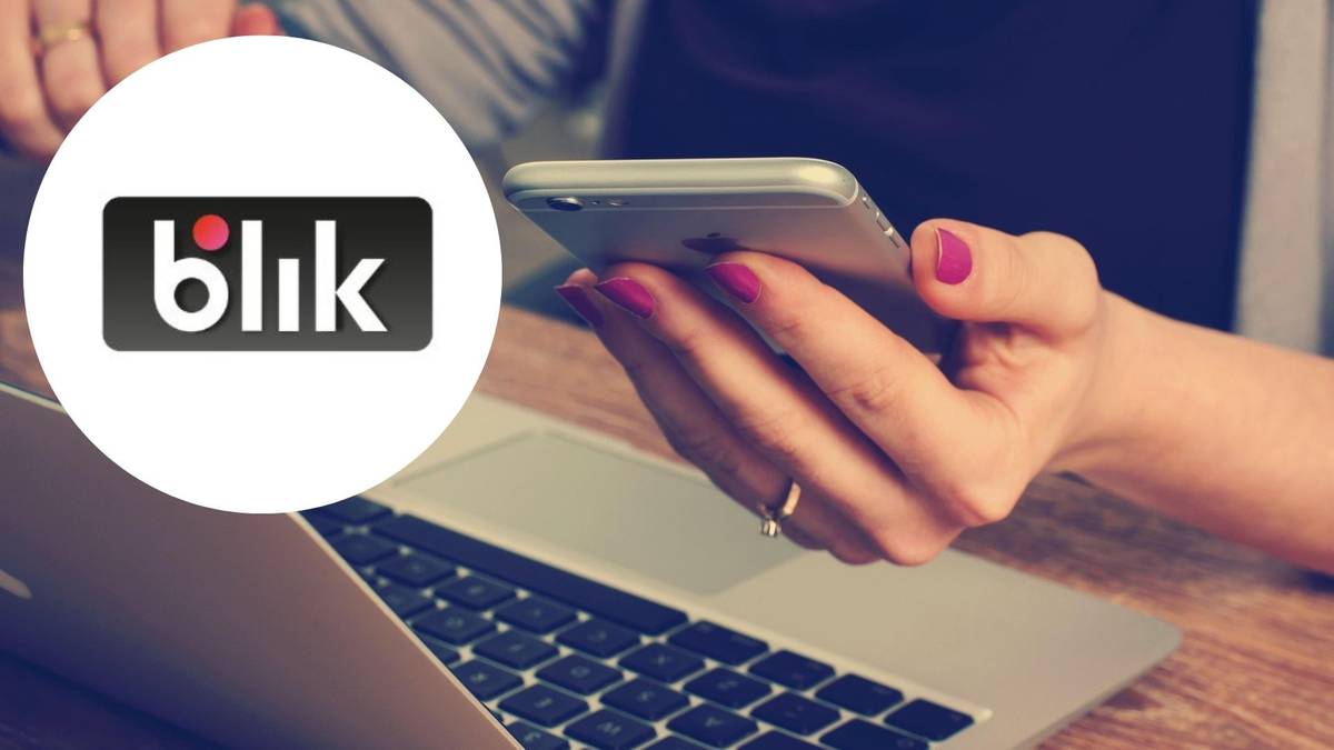 Logo systemu płatności Blik obok dłoni trzymającej smartfon na tle laptopa.