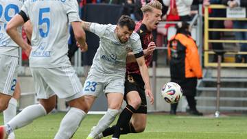 Dwóch piłkarzy walczących o piłkę na boisku podczas meczu, jeden z nich w jasnym stroju z numerem 23.