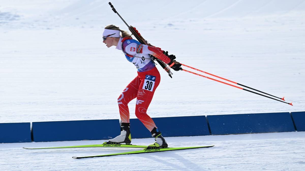 Biathlonistka w czerwonym stroju narciarskim z numerem 30 na plecach, z karabinem na plecach i kijami w rękach, biegnie na nartach po śniegu.
