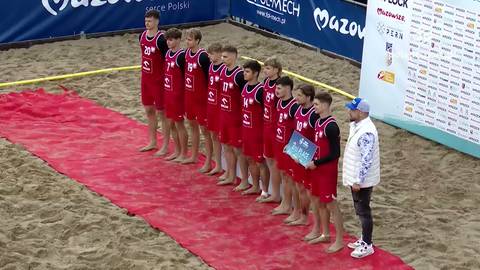 Drużyna chłopc&oacute;w w czerwonych strojach stoi w rzędzie na czerwonym dywanie na plaży podczas ceremonii wręczenia nagr&oacute;d.