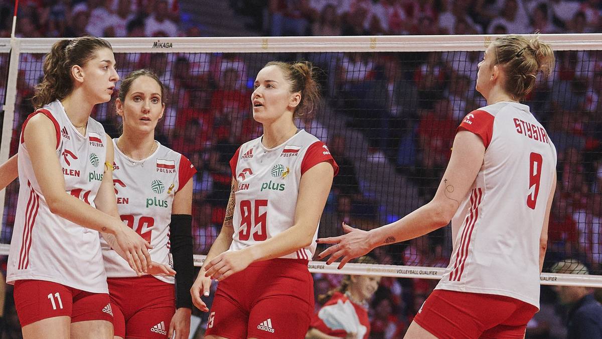 Cztery siatkarki w białych koszulkach z czerwonymi detalami i numerami na plecach stoją na boisku siatkarskim i rozmawiają ze sobą.