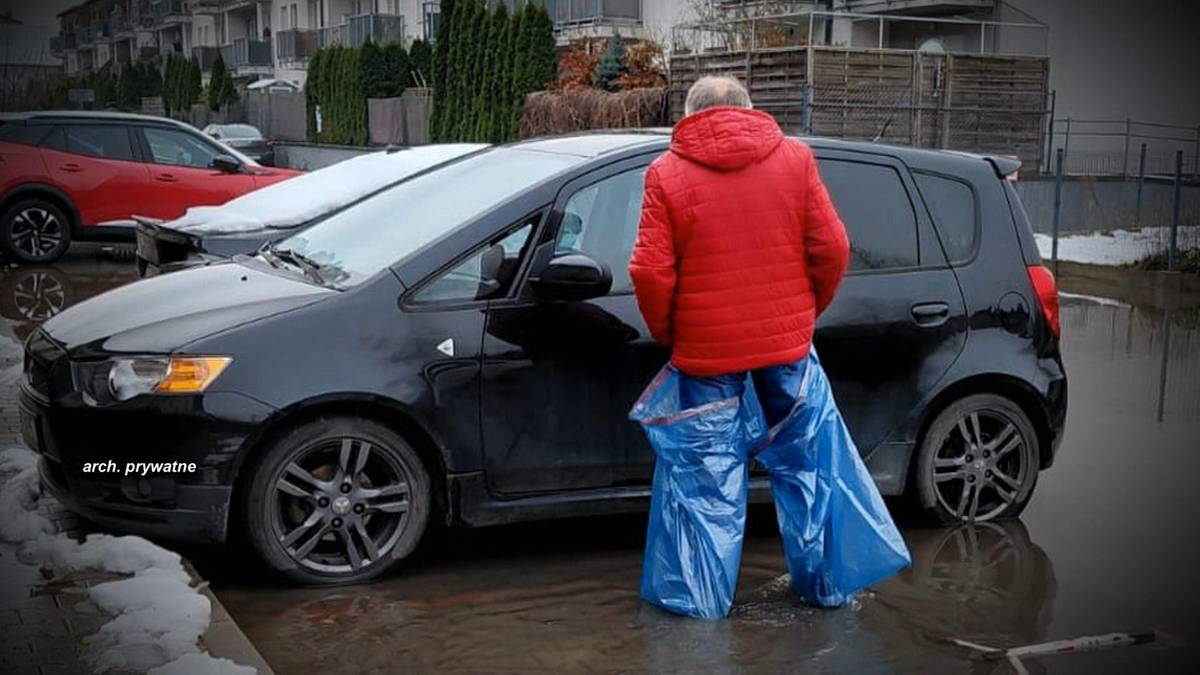 Mężczyzna w czerwonej kurtce zakłada worki na śmieci na nogi stojąc obok czarnego samochodu na zalanej ulicy.