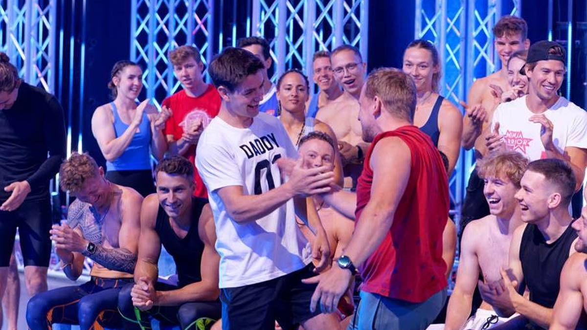 Grupa młodych mężczyzn w strojach sportowych pozuje do zdjęcia, niektórzy się uśmiechają. W tle widać niebieskie i białe światła.