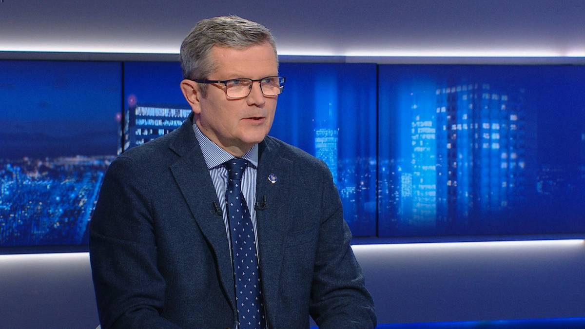 Mężczyzna w okularach i garniturze siedzi w studiu telewizyjnym.
