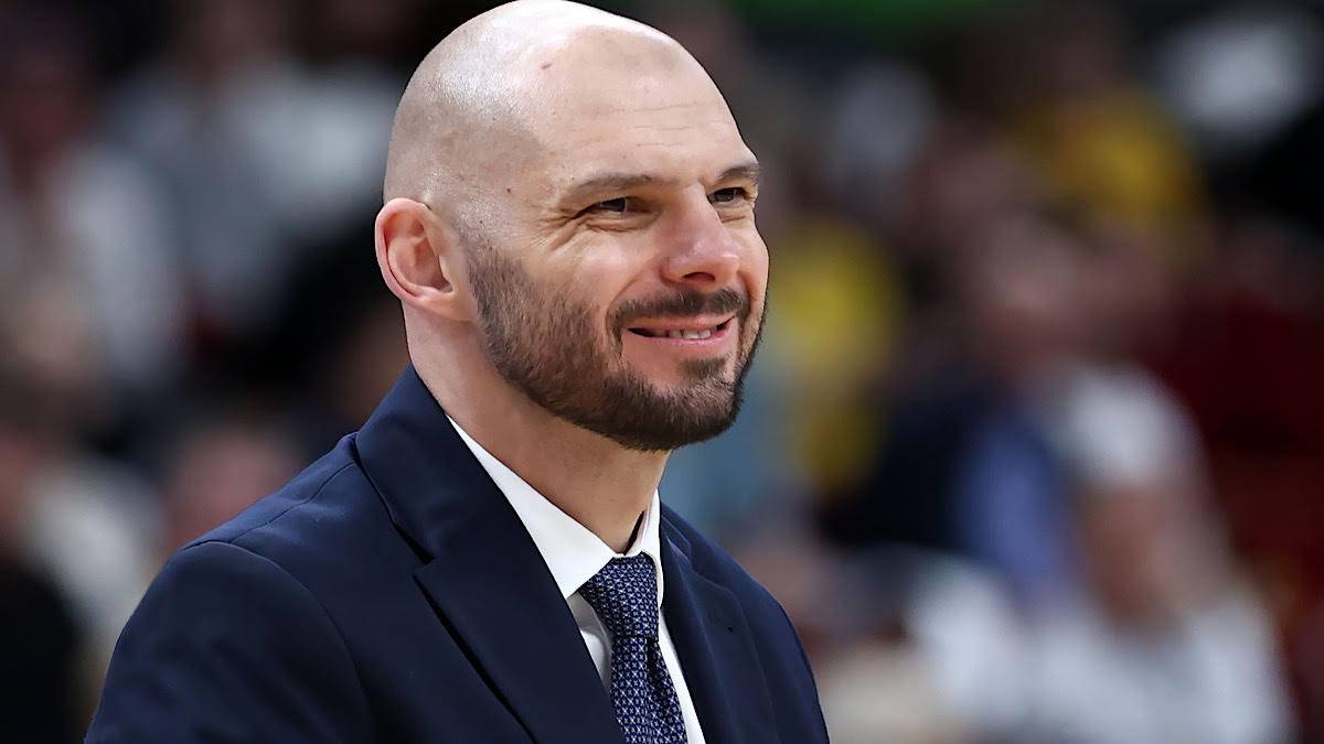 Portret mężczyzny w garniturze i krawacie, z ogoloną głową i zarostem, uśmiechającego się.