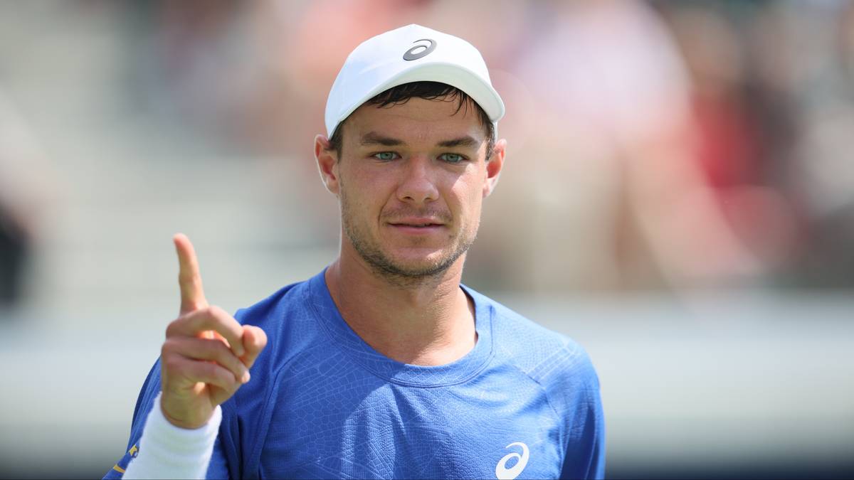 Mężczyzna w czapce z daszkiem i niebieskiej koszulce sportowej, podnosi palec wskazujący do góry.