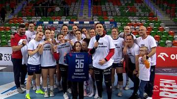 Grupa zawodniczek MKS Piotrcovii w strojach sportowych, z kt&oacute;rych jedna trzyma koszulkę z numerem 36, podczas gdy reszta stoi za nią, uśmiechając się do aparatu. W tle widać trybuny z widzami i elementy hali sportowej.