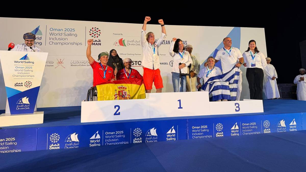 Trzech żeglarzy na podium, na środku zawodnik w białej koszulce podnosi ręce do góry, na pierwszym miejscu.