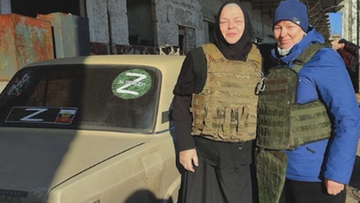 Dwie kobiety, jedna w stroju zakonnym i kamizelce taktycznej, druga w kurtce i czapce, pozują do zdjęcia obok samochodu z naklejkami "Z".