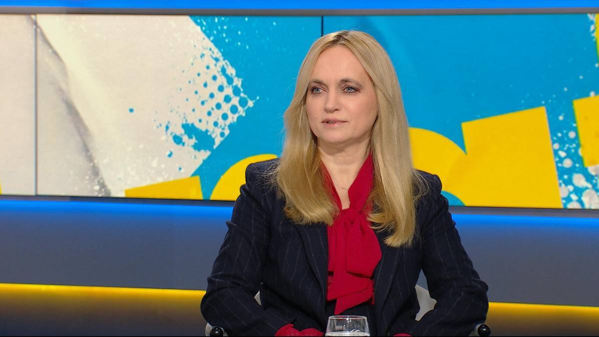 Kobieta w czerwonej bluzce i granatowej marynarce siedzi na krześle, w tle ekran z abstrakcyjnym wzorem.