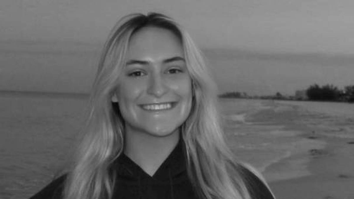 Portret młodej kobiety z długimi blond włosami, uśmiechającej się do kamery. W tle rozciąga się plaża i morze, a niebo jest zachmurzone.
